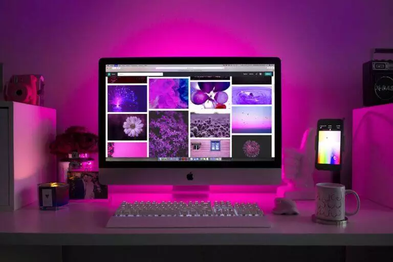 A desktop computer displaying purple-themed images is on a desk with a glowing keyboard, mouse, smartphone, mug, candle, and decor, illuminated by purple lighting. This workspace perfectly complements any WordPress development environment focused on creativity and productivity.